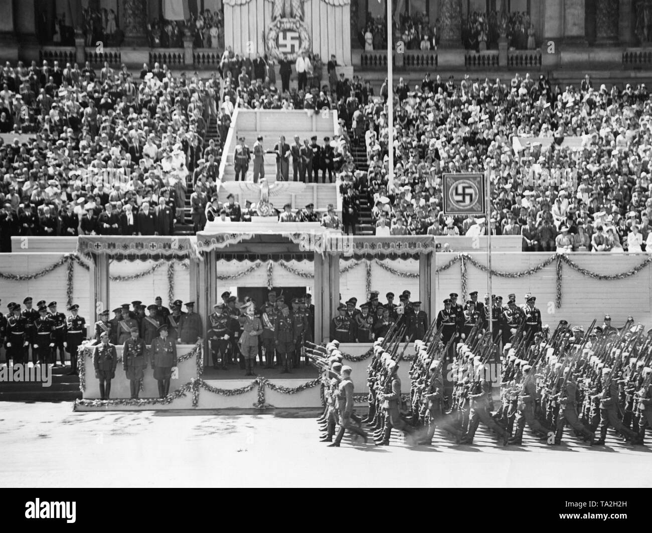 Photo of the VIP stand during the parade for the Condor Legion on the East-West Axis (former Chalottenburger Chaussee, today Strasse des 17. Juni) in front of the main facade of the Technische Universitaet in Berlin on June 6th, 1939. Adolf Hitler (under a canopy) is giving the marching soldiers the Nazi salute. On the left under the Reichsadler (imperial eagle) with a swastika, a radio commentator. Stock Photo