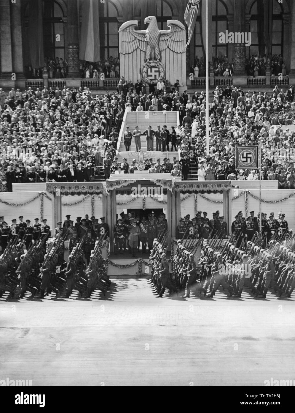 Photo of the VIP stand during the parade for the Condor Legion on the East-West Axis (former Chalottenburger Chaussee, today Strasse des 17. Juni) in front of the main facade of the Technische Universitaet in Berlin on June 6th, 1939. Adolf Hitler (under a canopy) is giving the marching soldiers the Nazi salute. In the center under the Reichsadler (imperial eagle) with a swastika, a radio commentator. Stock Photo