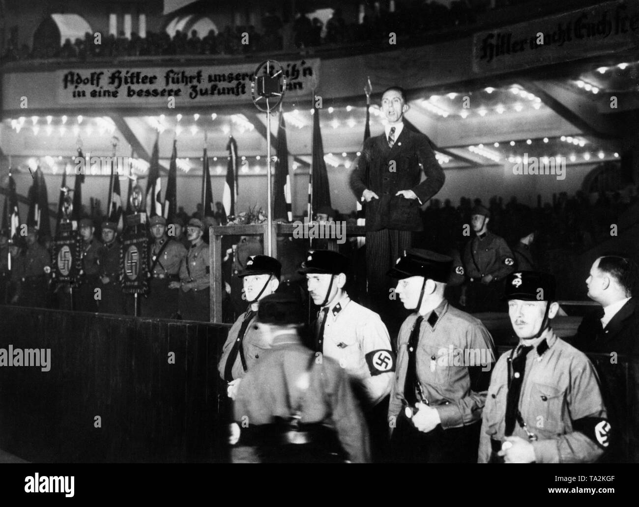 Joseph Goebbels holds a campaign speech at the Berlin Sports Palace during the Reichstag election campaign of 1932. Before him stands a group of SS men as personal stewards. The slogans on the banners read: 'Adolf Hitler leads our ranks into a better future ...' and 'Hitler makes you free'. Stock Photo