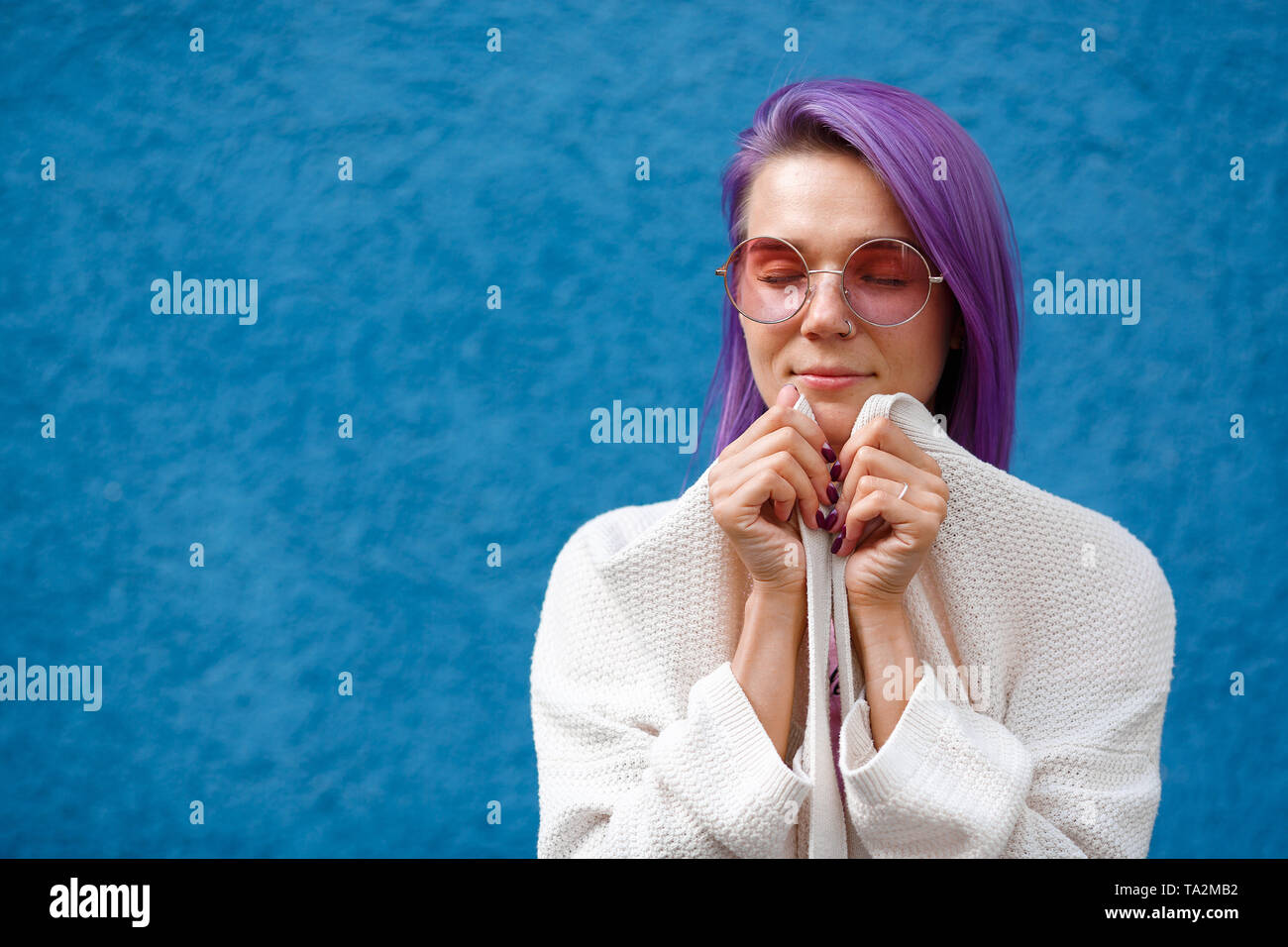 a girl with violet hair on a blue background closed eyes, wearing pink glasses, wrapped in a jacket, smiling, calm, beautiful hands, bit cold Stock Photo