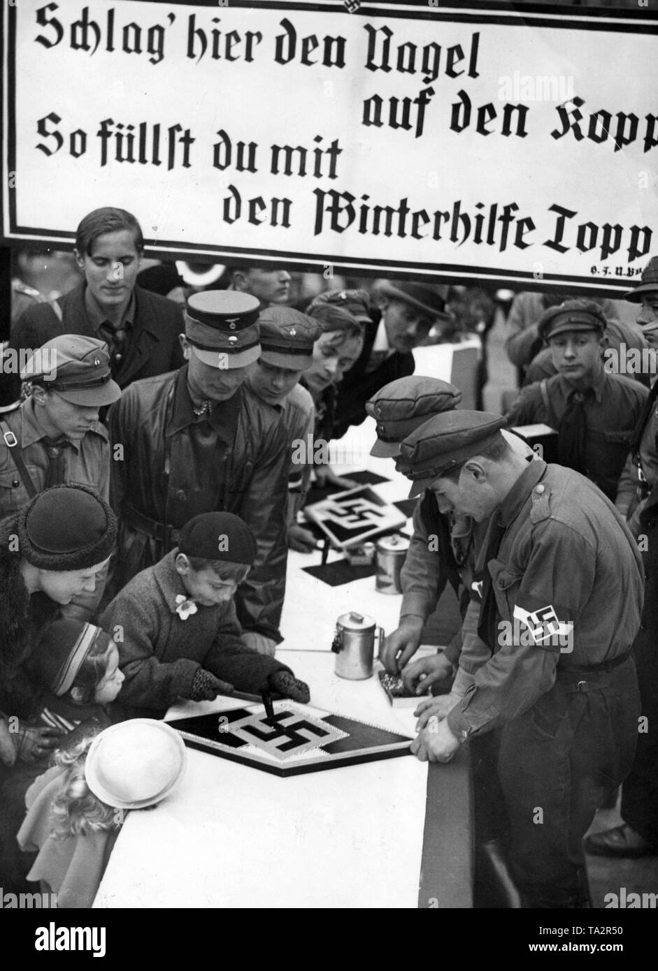 Children are hitting nails into a coat of arms of the Hitler Youth (HJ) during a donation campaign for the Nazi Winter Relief. Stock Photo