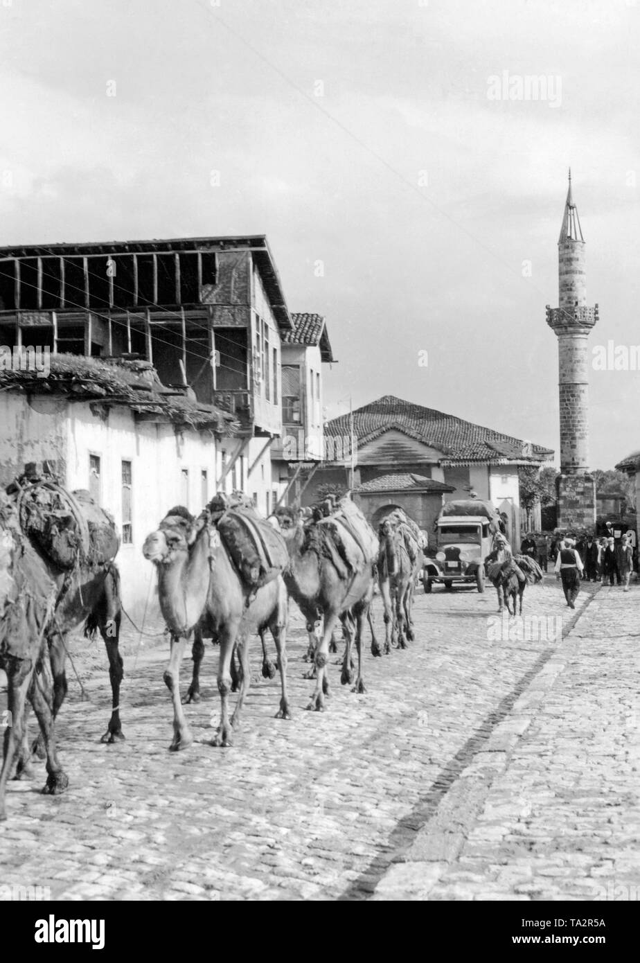 Street life in a city in central Turkey - in the background a mosque, on the road next to a loaded car a caravan of camels. Stock Photo