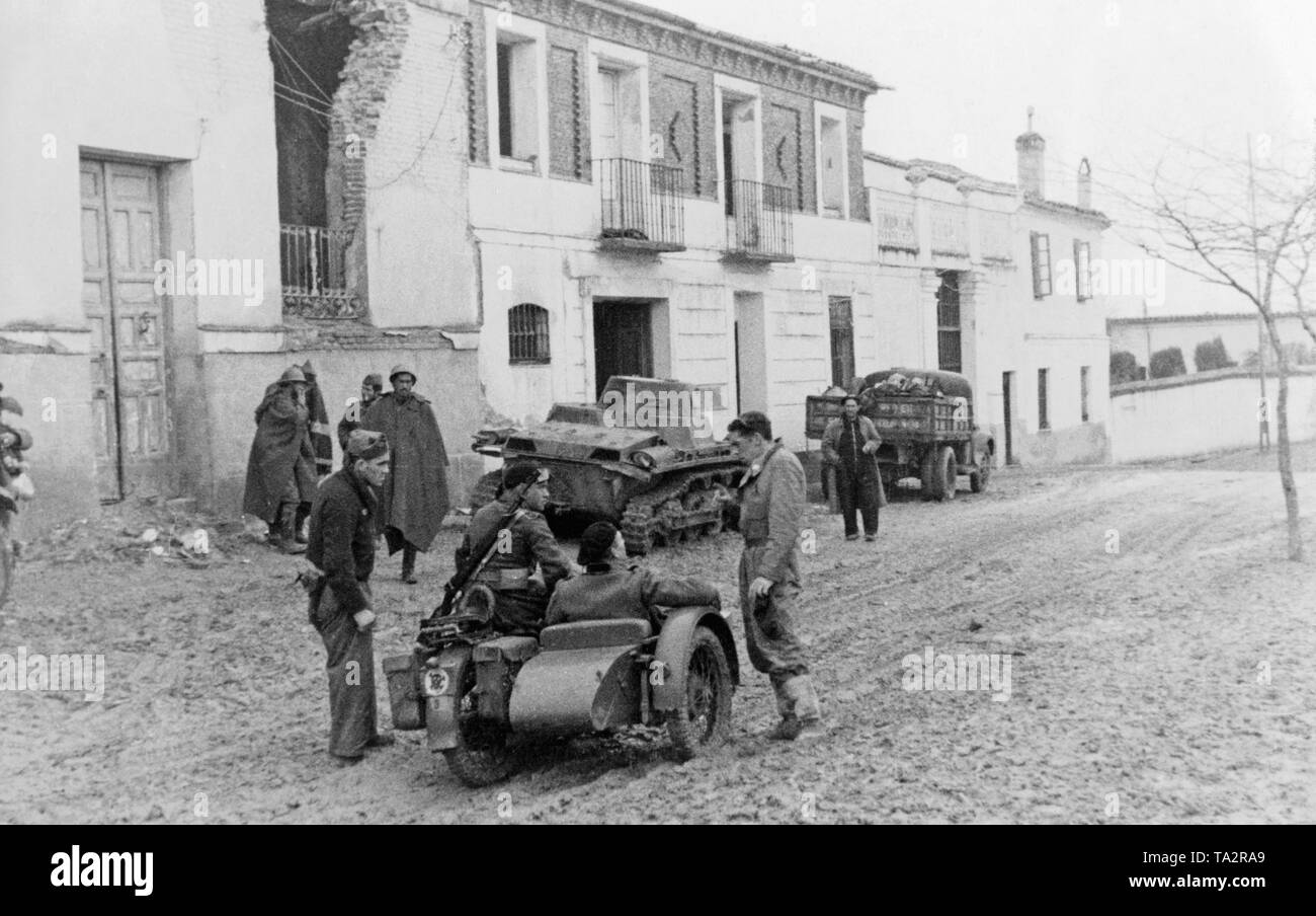 After the conquest of Malaga and Mortil on February 8, 1937, Spanish national motorized units under General Gonzalo Queipo de Llanos continued their march towards Almeria. In the foreground, a motorcycle with a sidecar. In the background, a German armored fighting vehicle I (PzKpfw I) and a truck that is surrounded by soldiers. In the background, a partially destroyed street. Stock Photo