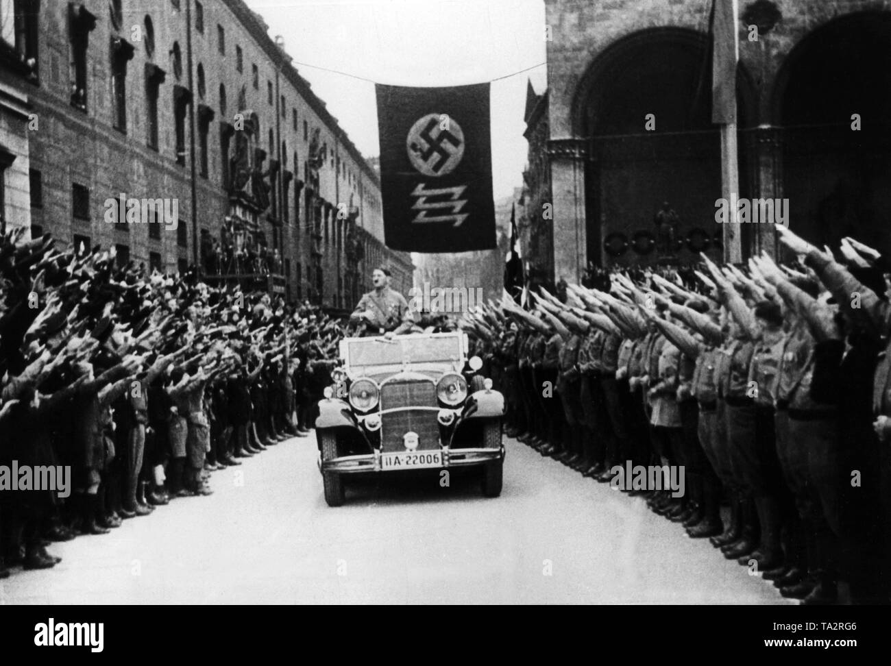 Adolf Hitler drives in an open car along the Residenzstrasse in Munich. To the right and left of the street is the Feldherrenhalle and the Residenz. Between the two buildings is a flag with swastika and a triple siegrune. Undated photo. Stock Photo