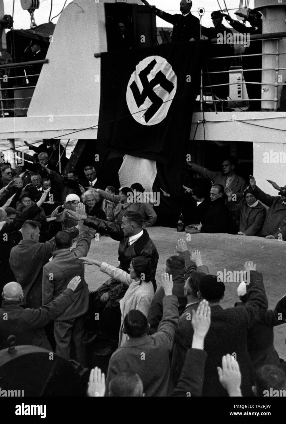 Holidaymakers of the Nazi organization 'Kraft durch Freude' show the Nazi salute aboard a Kdf-ship. Above a swastika flag a ship officer. Stock Photo