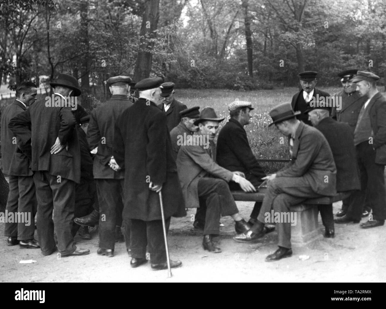 Some Men Sit On A Park Bench And Play Cards Others Stand Around