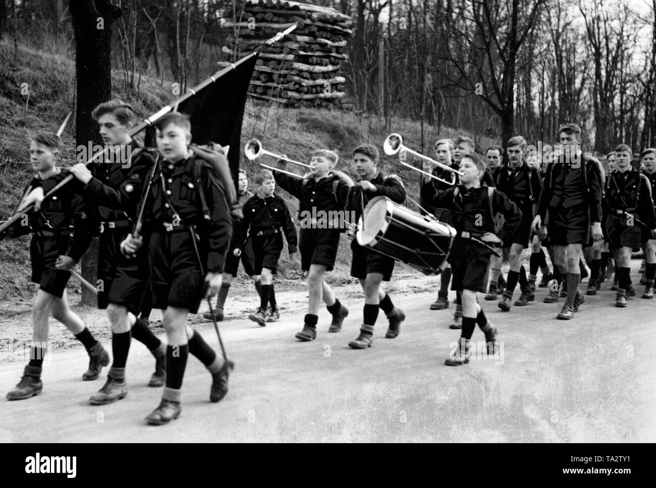 Pimpfs are marching in line with music along a forest path. In the background, a pile of stere wood. Stock Photo