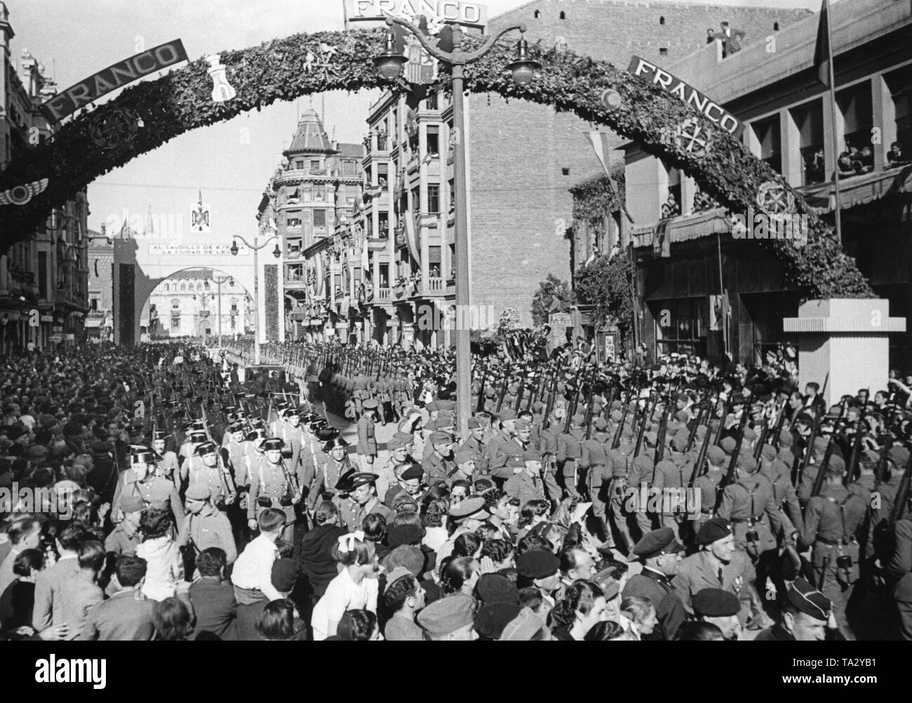 Photo of a victory parade of Spanish national troops and the German Condor Legion in honor of General Francisco Franco in the festively decorated streets of Ciudad de Leon, Castile and Leon on May 22, 1939. The soldiers are marching in two directions: on the left a unit of the Spanish Guardia Civil is parading, on the right, the Condor Legion. In the background, the triumphal arch in honor of the Condor Legion. Stock Photo