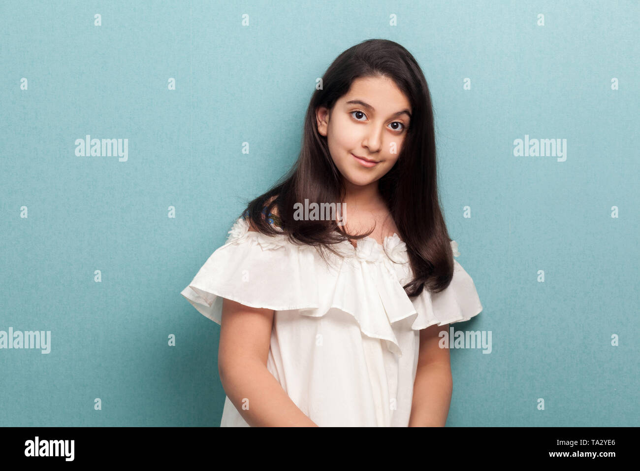 Portrait of happy calm beautiful brunette young girl with black long straight hair in white dress standing and looking at camera with smile. indoor st Stock Photo