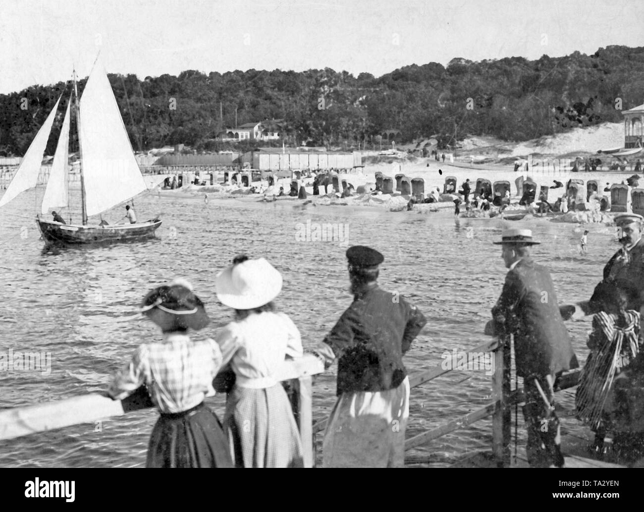 Vacationers look at a sailboat from the jetty. On the beach are beach chairs that protect the bathers from the sun. Stock Photo