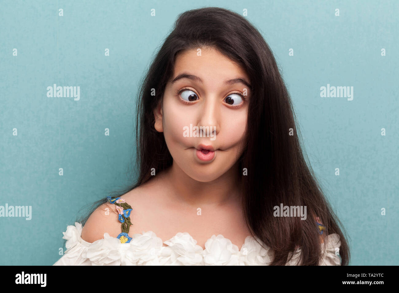 Portrait of funny beautiful brunette young girl with black long straight hair in white dress standing with crossed eyes, fish lips and crazy expressio Stock Photo