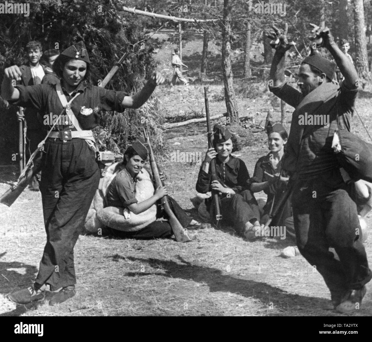 Photo of two dancing Republican fighters behind the front line in Sierra Peguerinos northwest of Madrid. The dancer on the left is dressed in overall and is armed with a Gewehr 98 rifle. She is wearing the chevron of a Teniente (lieutenant) on her cap. Her dancing partner plays the castanets and wears a blanket and carries a bag. In the background, fighters and female fighters watch amused. Stock Photo