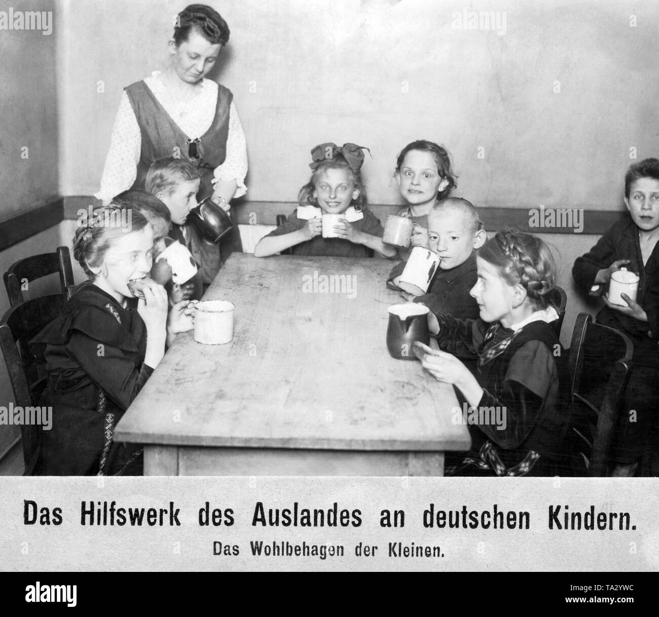 Eight children sit in a room - most around a table - each with a cup and  something to eat. On the left stands a woman who gives a boy a drink