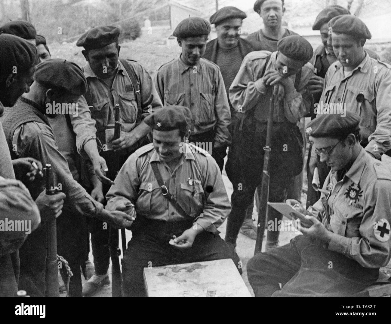 A lieutenant (with a star on his cap) of a volunteer unit of the Spanish national troops, pays his soldiers while sitting at a table (in breeches and field tunic). Behind him on the left, a corporal (three strips on his chest). On the right side of the table, there is a military doctor sitting  with a crab badge, and he is taking notes of the payouts. On the table, a pile of silver coins. Stock Photo