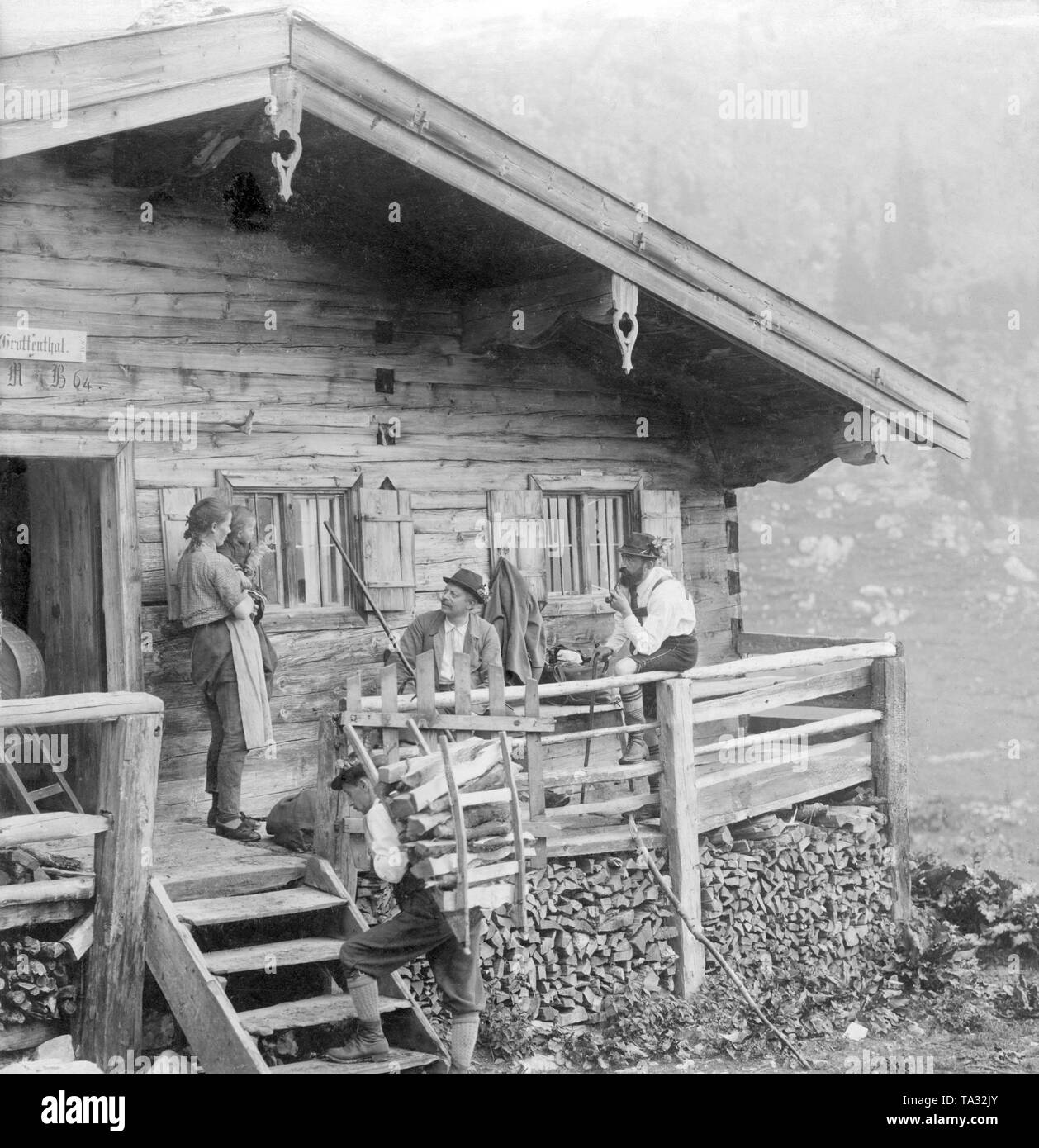 The dairymaid (left) with her child in her arms greets the first two 'seasonal guests' (right) on an alpine pasture in Upper Bavaria. In the foreground, a helper of the dairymaid carries wood for the stove into the house on his back. Stock Photo