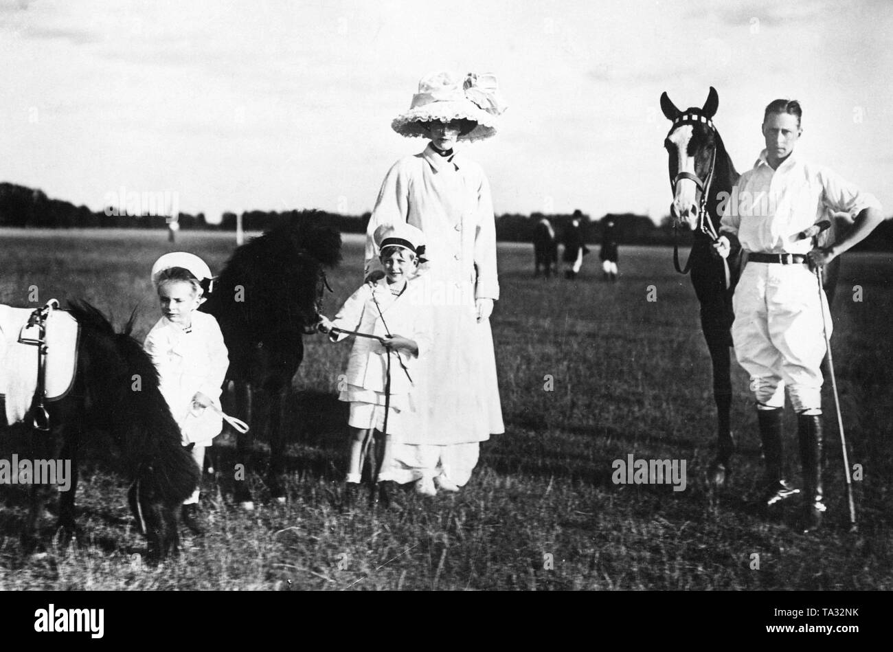 From left to right: Prince Louis Ferdinand, Prince William, Crown Prince Cecilie, Crown Prince William.  The picture was made probably during a polo game of the Crown Prince. The two children steady their riding ponies while Crown Prince Wilhelm is dressed as a polo player and stands with a racket in his hand next to his horse. Stock Photo