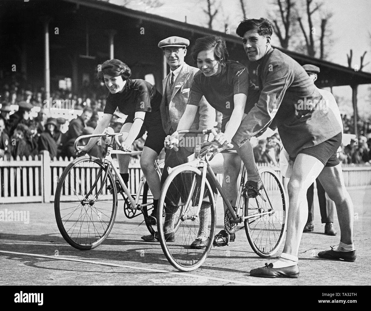 Two young track cyclists wait for the start of a race on April 1, 1929. They and their bikes are supported by helpers. In the background, spectator stands. Stock Photo