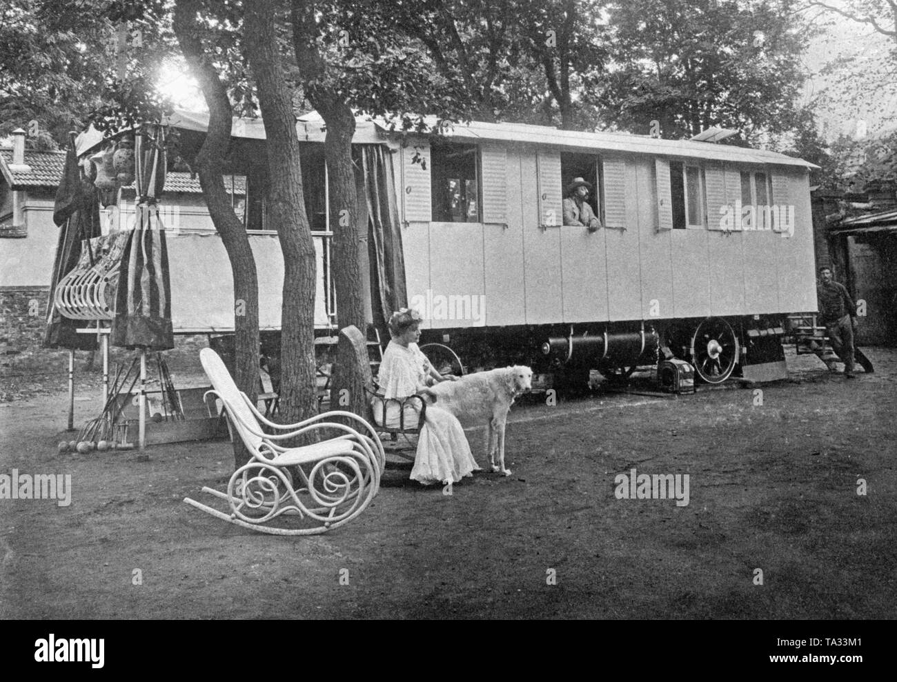 Historical, luxurious motorhome ( 'Motor Touring Car'), in front of it a woman is sitting in a rocking chair, beside her is a dog. A man stands at a window of the caravan and is looking out. Stock Photo