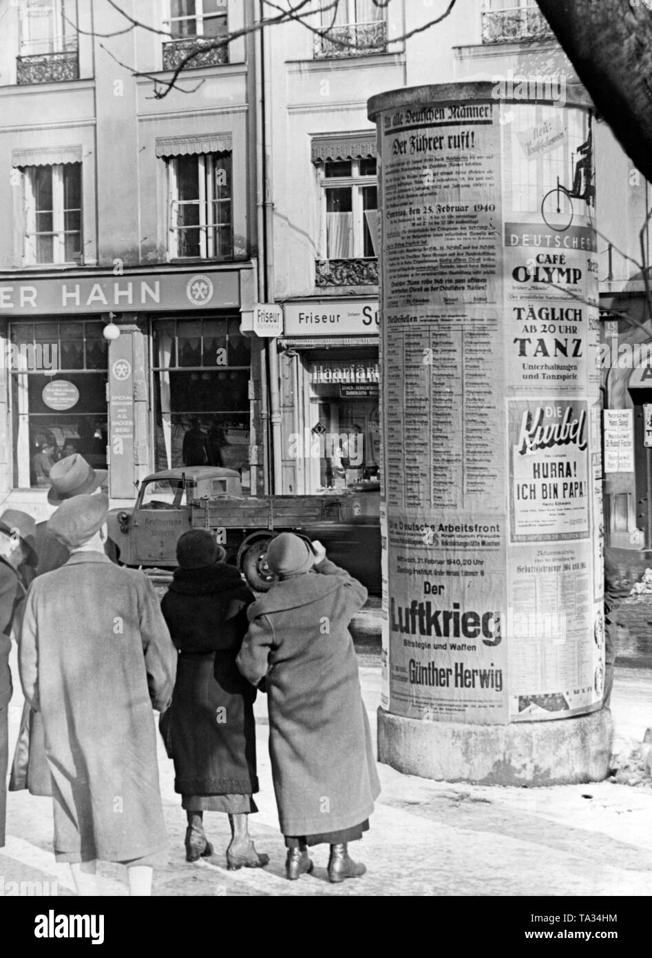 Passers-by observe the propaganda posters on an advertising column in a Bavarian city, probably Munich. A propaganda poster is entitled 'Der Fuehrer ruft' ('The Fuehrer is calling') and declares the mobilization of the year 1922. Another poster of the German Labor Front (DAF) announces the lecture of Guenther Herwig 'Der Luftkrieg Strategie und Waffen' ('The air war strategy and weapons'). Stock Photo