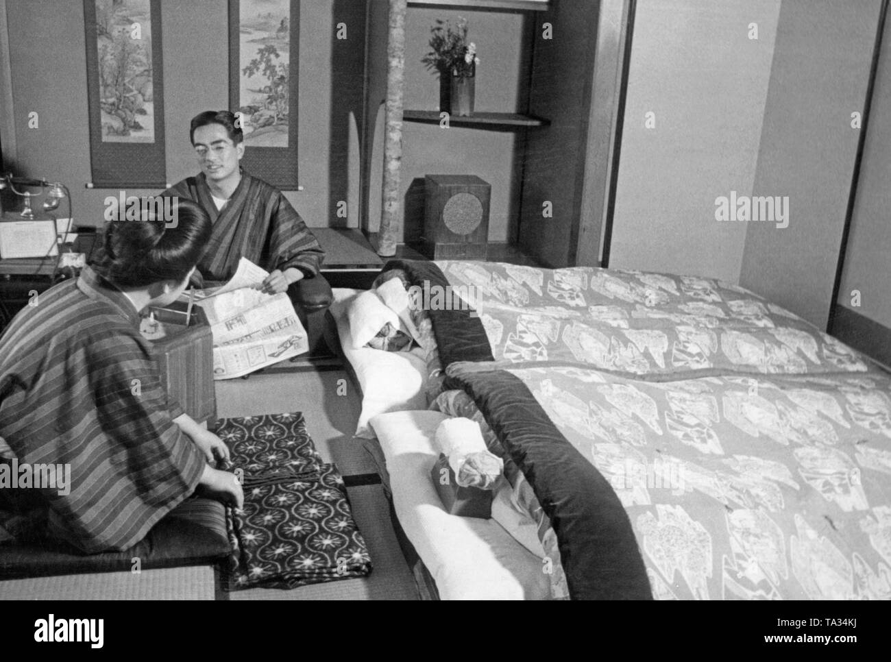A man and a woman in kimonos sit on the floor of their living room and talk. The man is holding a newspaper in his hand, their beds are next to them. Stock Photo