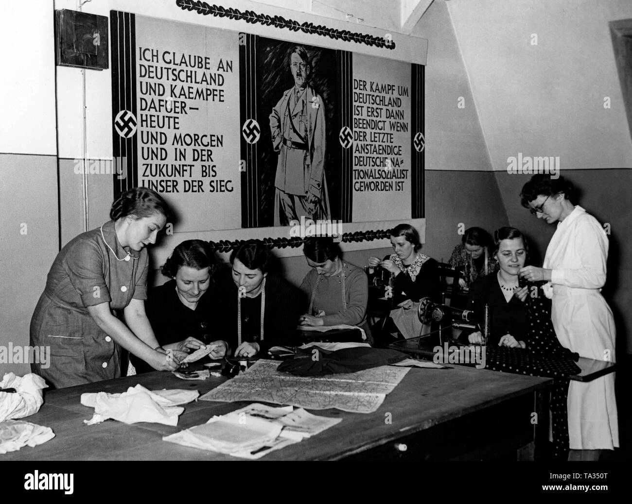 Photo of the participants of a sewing course of the Reichsmuetterdienst. On the left, the social worker, who guides the other women. In the background a propaganda poster with Adolf Hitler and the inscription 'I believe in Germany and fight for it - today and tomorrow and in the future until we win. The fight for Germany is only finished, when the last decent German has become a National Socialist'. Photo of the Propaganda Company (PK) Stock Photo
