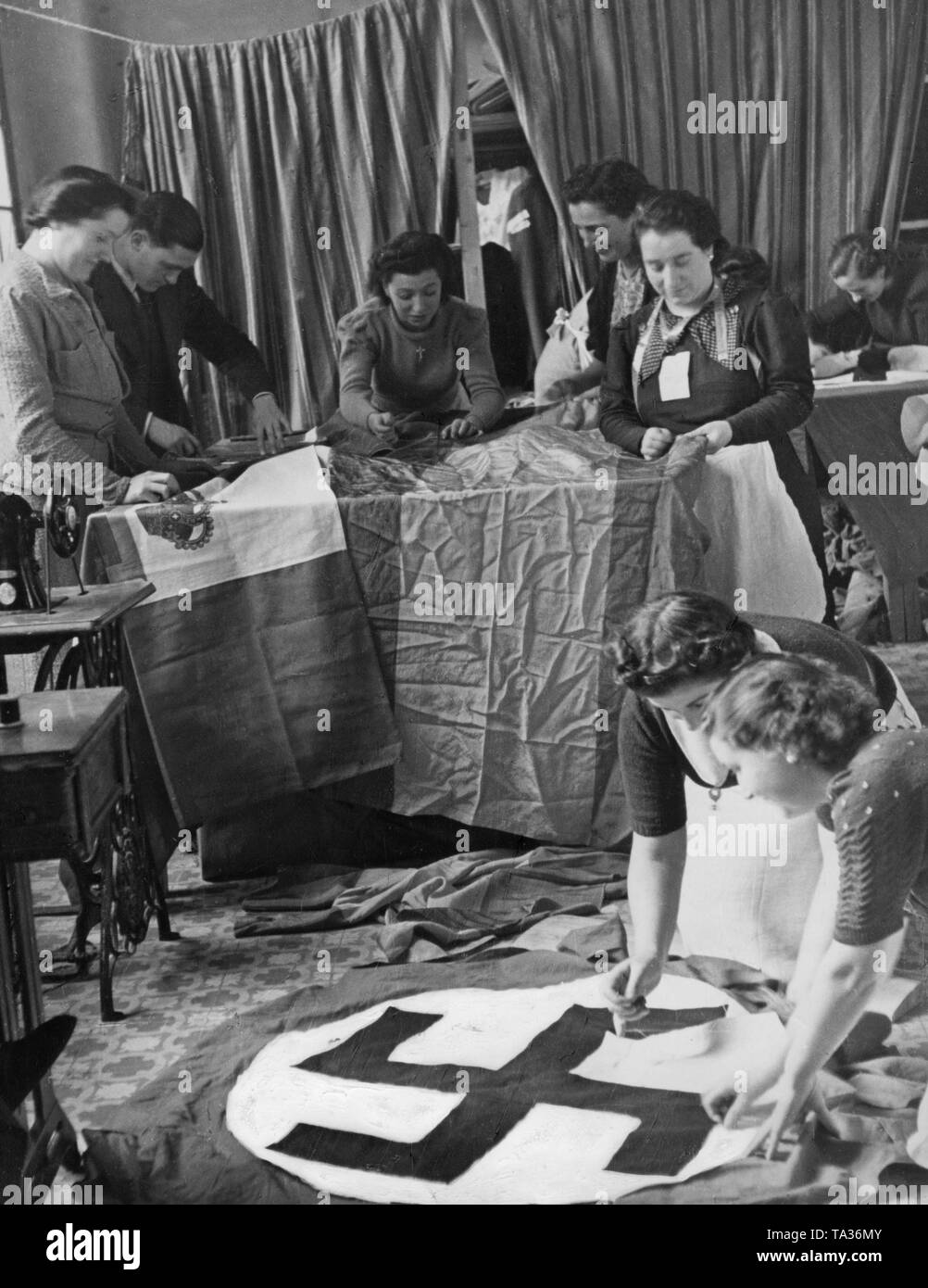 Undated photo of a group of Spanish women sewing Spanish, Italian and German flags for the Condor Legion in a workshop. In the foreground two women are sewing a swastika onto a flag. On the table, two banderas are being sewn. Stock Photo