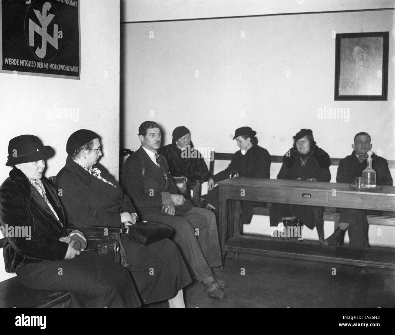 Several women and two men are waiting in the antechamber of a women's counseling center of the German Labor Front in Kaiser-Wilhelm-Strasse (now Karl-Liebknecht-Strasse), Berlin. On the left, a promotional poster of the NS-Volkswohlfahrt: 'Become a member of the NS-Volkswohlfahrt'. To the right on the wall, a portrait of Adolf Hitler. Stock Photo