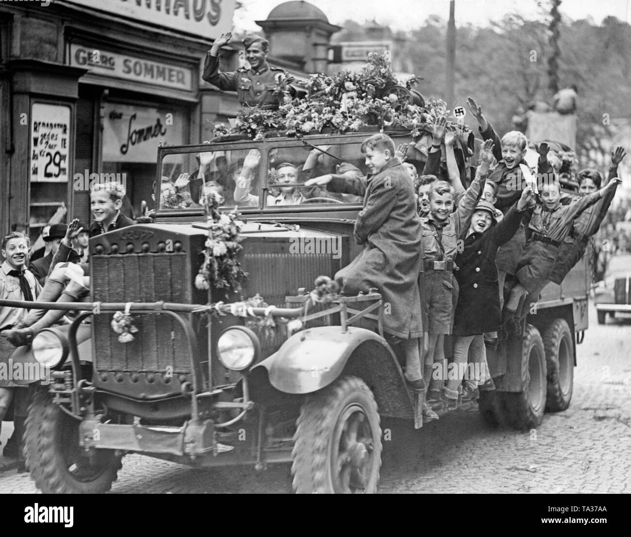 People receive the retreating German troops in Karlsbad (today Karlovy Vary) on October 4, 1938. Some children have climbed on a truck, and accompany them. The truck is decorated with flowers. Stock Photo