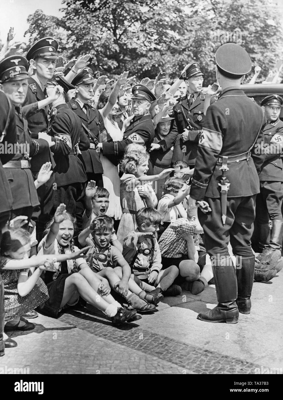Photo of a group of enthusiastic spectators on Unter den Linden Strasse in Berlin, who are giving the Hitler salute to the marching troops of the Condor Legion on the occasion of their return from Spain on June 6, 1939. They are held back by SS men. Children are sitting on the pavement. Stock Photo