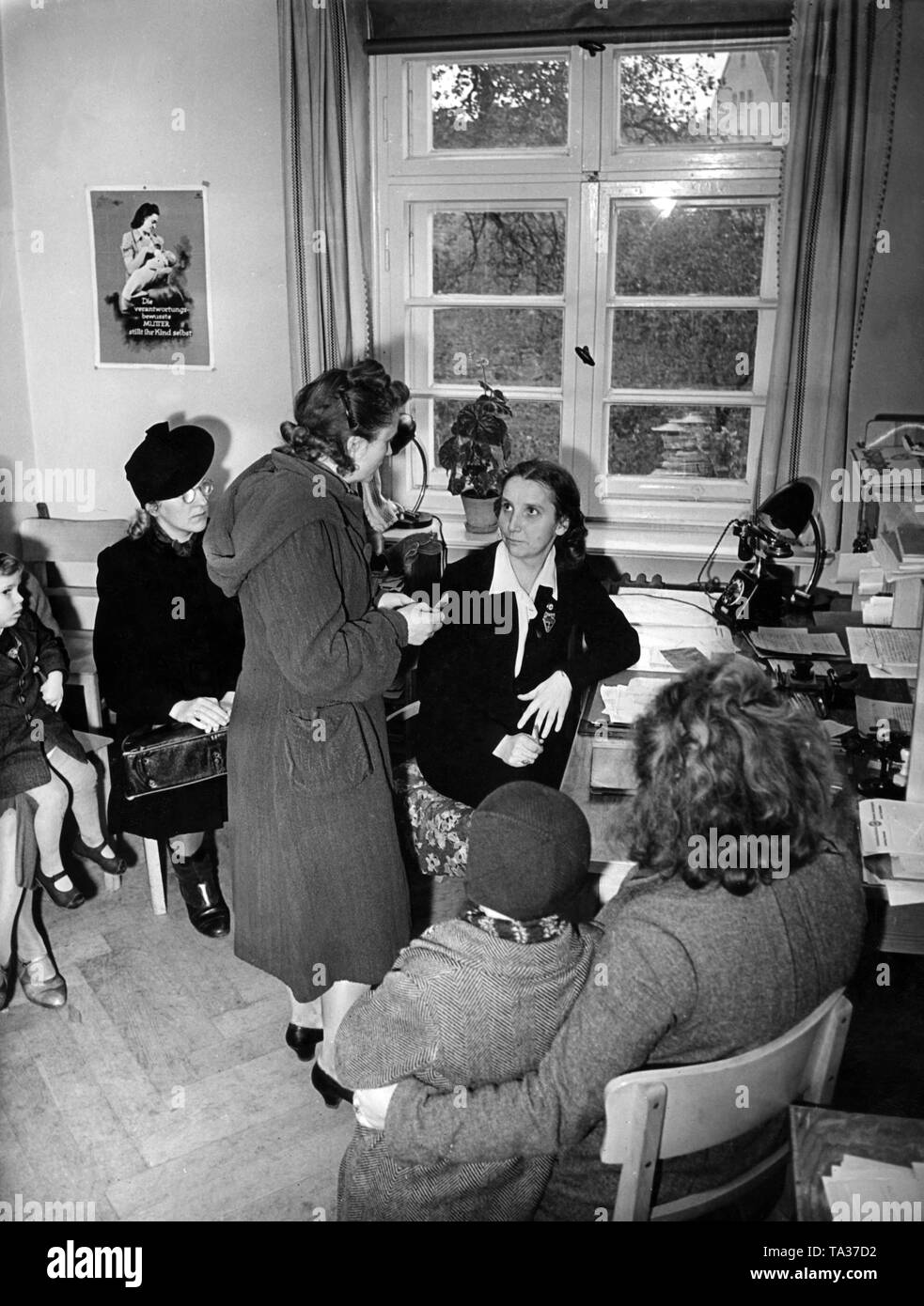 The leader of a local group of the NS-Frauenschaft helps women evacuated before the bomb war with advice and assistance regarding accomodation. On the left, a poster with a mother and child, as well as the lettering 'The responsible mother nurses her child herself'. Stock Photo