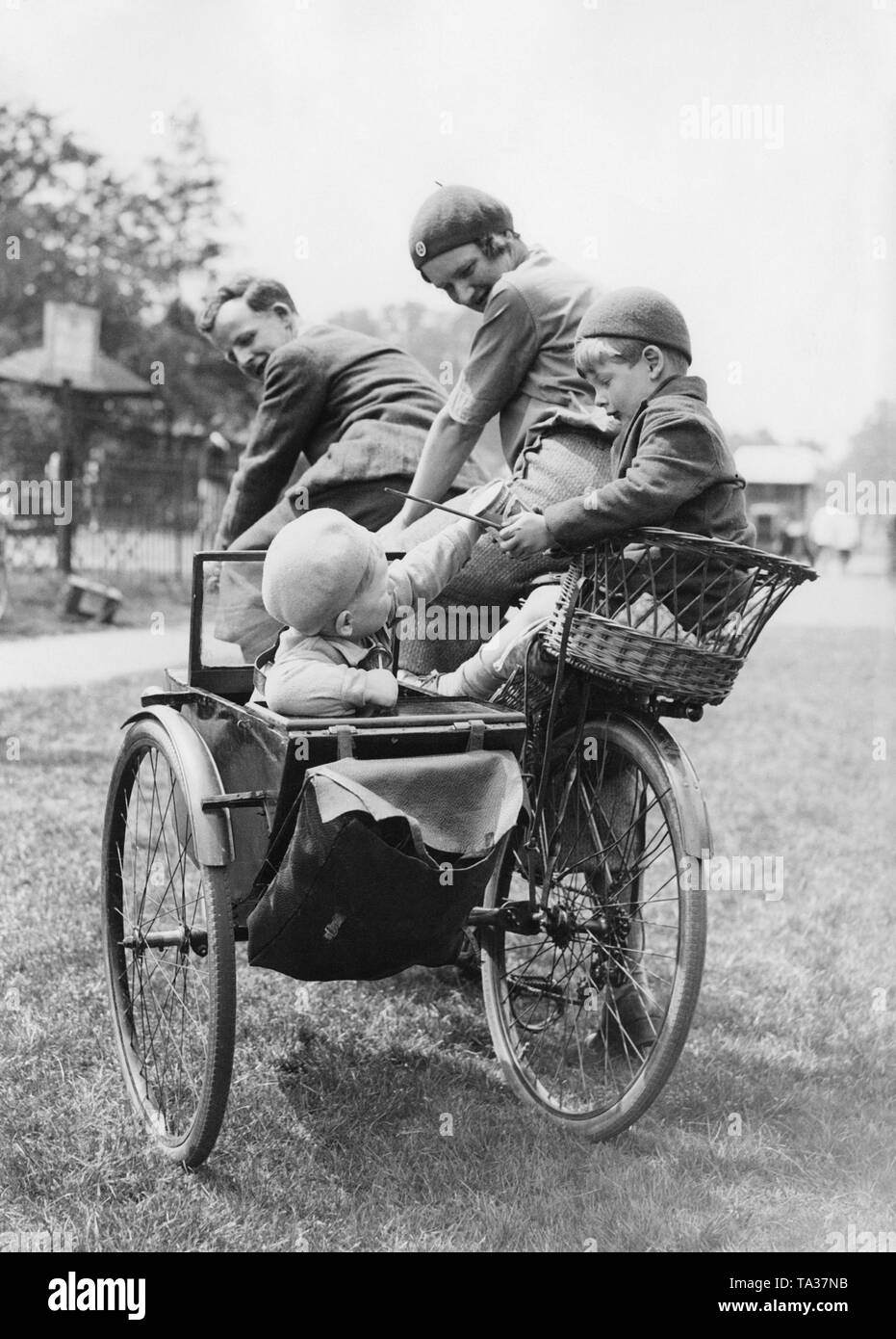 A family with two children is riding a tandem with sidecar through a park. The parents look at their two playing children. The older boy holds a can and a stick in his hand, while the younger child reaches for it. Stock Photo