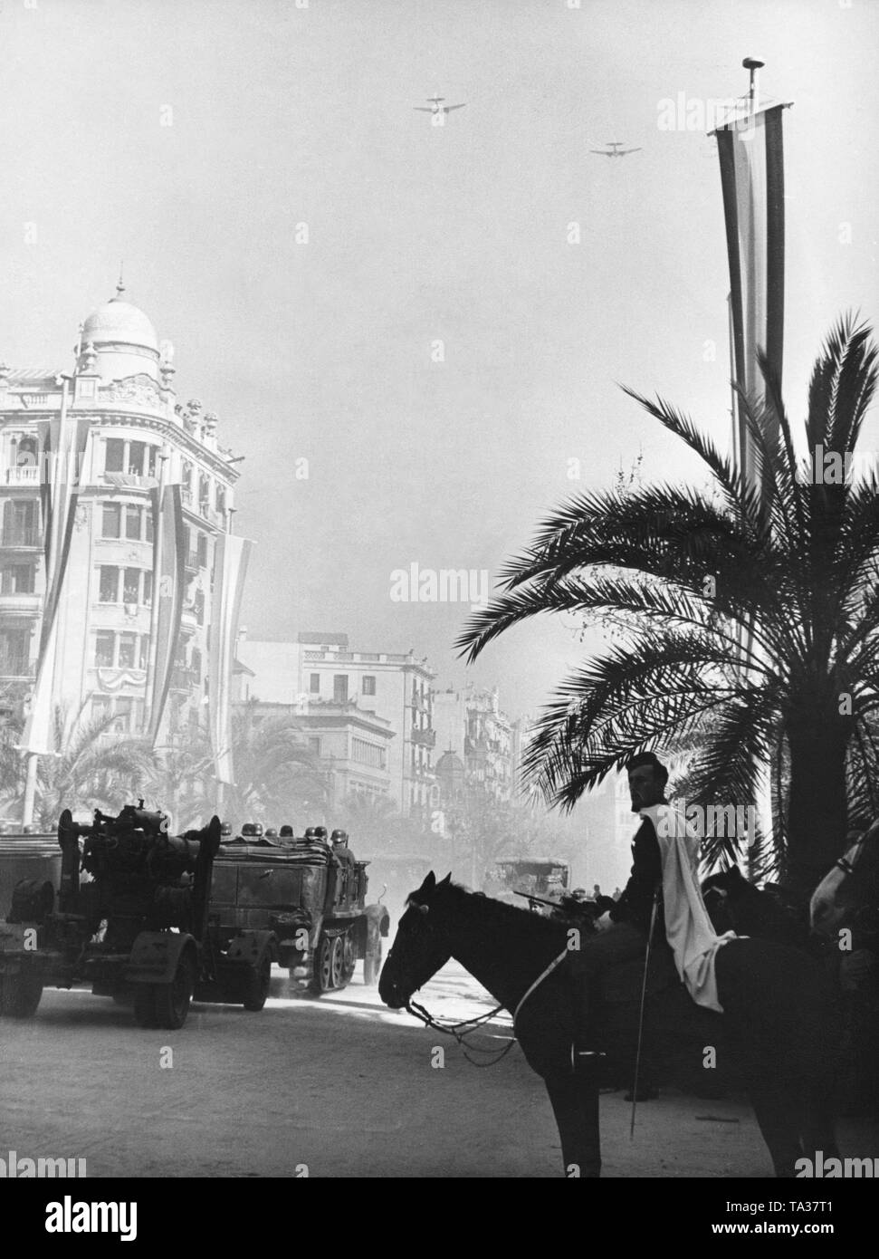 Photo of a victory parade of Spanish national units in Passeig de Colon after the conquest (January 1939) of Barcelona by General Francisco Franco in February, 1939. A German artillery procession of the Condor Legion is passing by. The 8.8cm FLAK 36 (two-axle mount) is carried by a heavy half-track vehicle, type 8 and 9. In the sky, formation flight of the German Condor Legion. Stock Photo