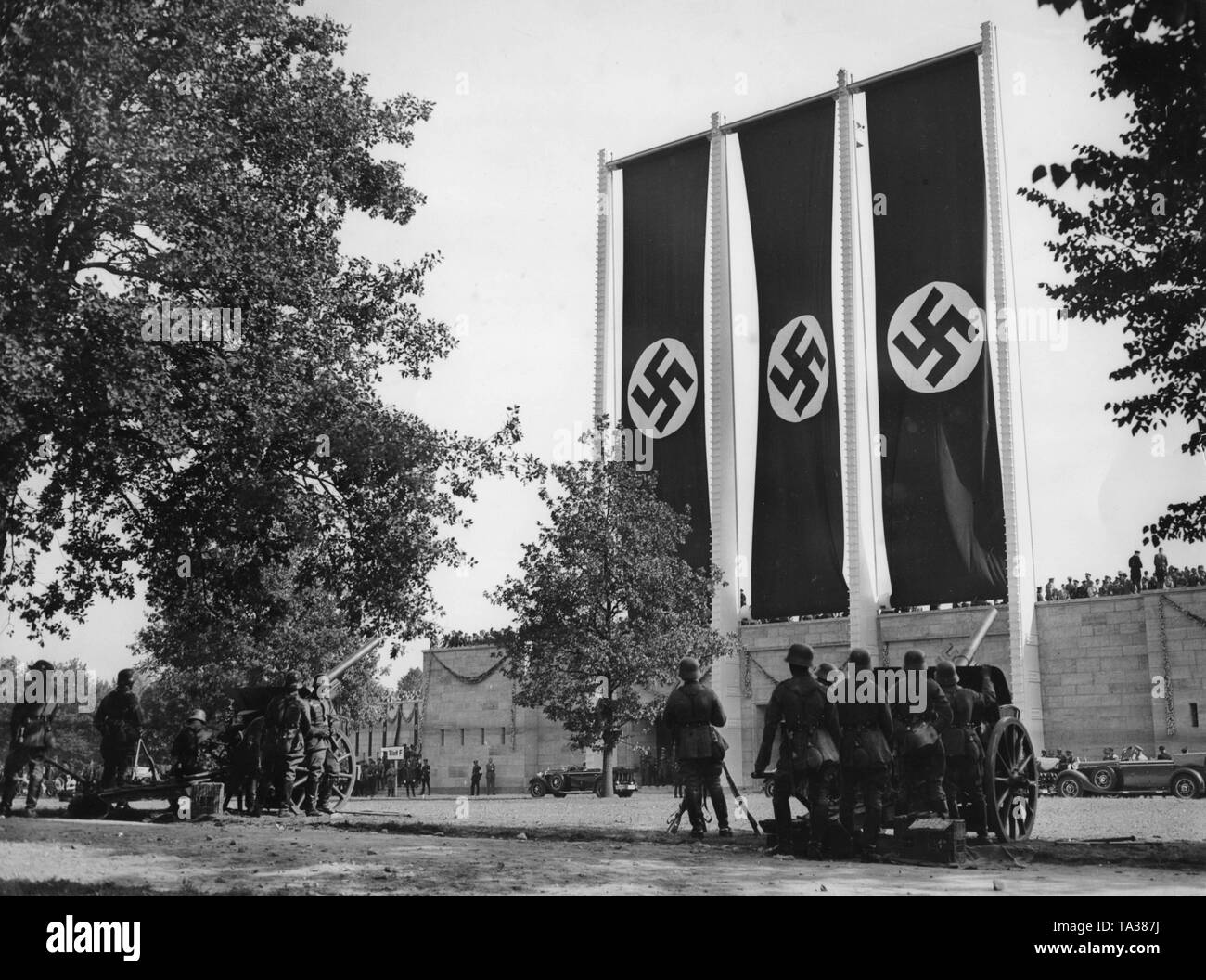 Units of the Reichswehr are giving cannon salutes at the muster of the SA and SS at the Nuremberg Rally behind the Luitpolt Arena with three oversized swastika flags. Stock Photo