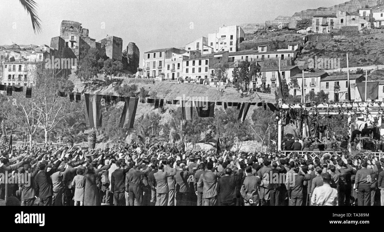 Photo of a crowd greeting the marching-in Francisco de Borbon y de la Torre, Duke of Seville (in the back riding a horse, from 1882 to 1953) in front of the ruins of the Castillo Gibralfaro in Malaga, Andalucia, Spain, in 1936. In the foreground, passers-by greet him. The street is decorated with Spanish flags. Stock Photo