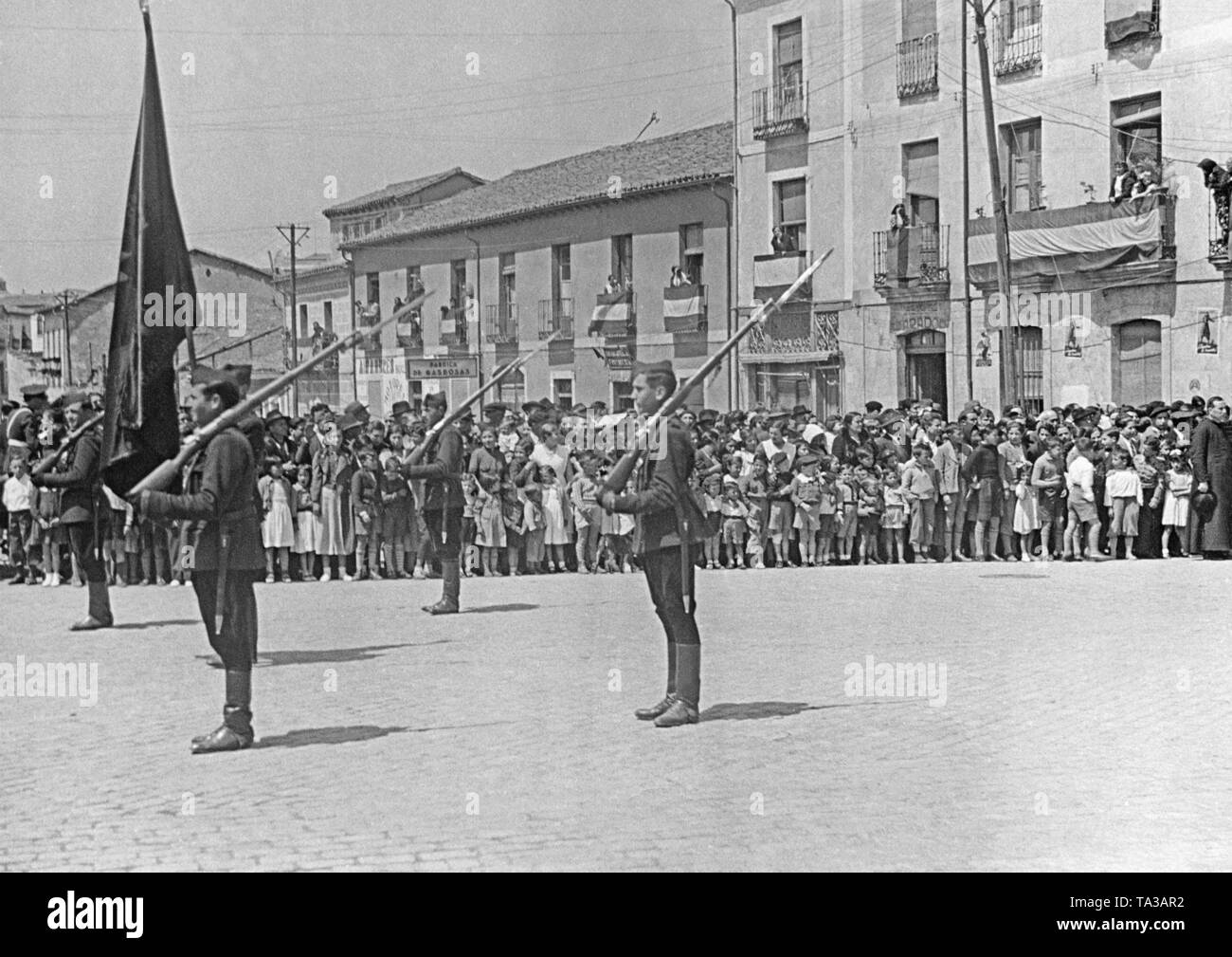 Photo of the main square of Valmaseda (30km south-east from Bilbao), Basque Country, Spain, in the summer of 1937. The red-black-red flag (left) of the Falange Espanola (Fascist Party of Spain), with yoke and arrows, is surrounded by four guards. In the background, villagers and children are watching. A few days earlier, Bilbao was conquered and fell prey to the Spanish national faction in June, 1937. Stock Photo