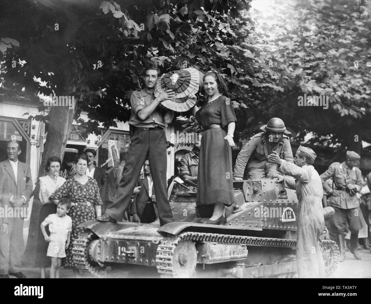 Photo of an Italian armored car (L3 / 33, Carro veloce 33) in the streets of Santander on August 27, 1937. A man and a woman are standing on the tank with a rosette decorated with national colors. Behind, a tank commander of the Italian Corpo Truppe Volontarie (CTV) leans out of his vehicle. In the background on the right, a German officer of the Condor Legion. Stock Photo