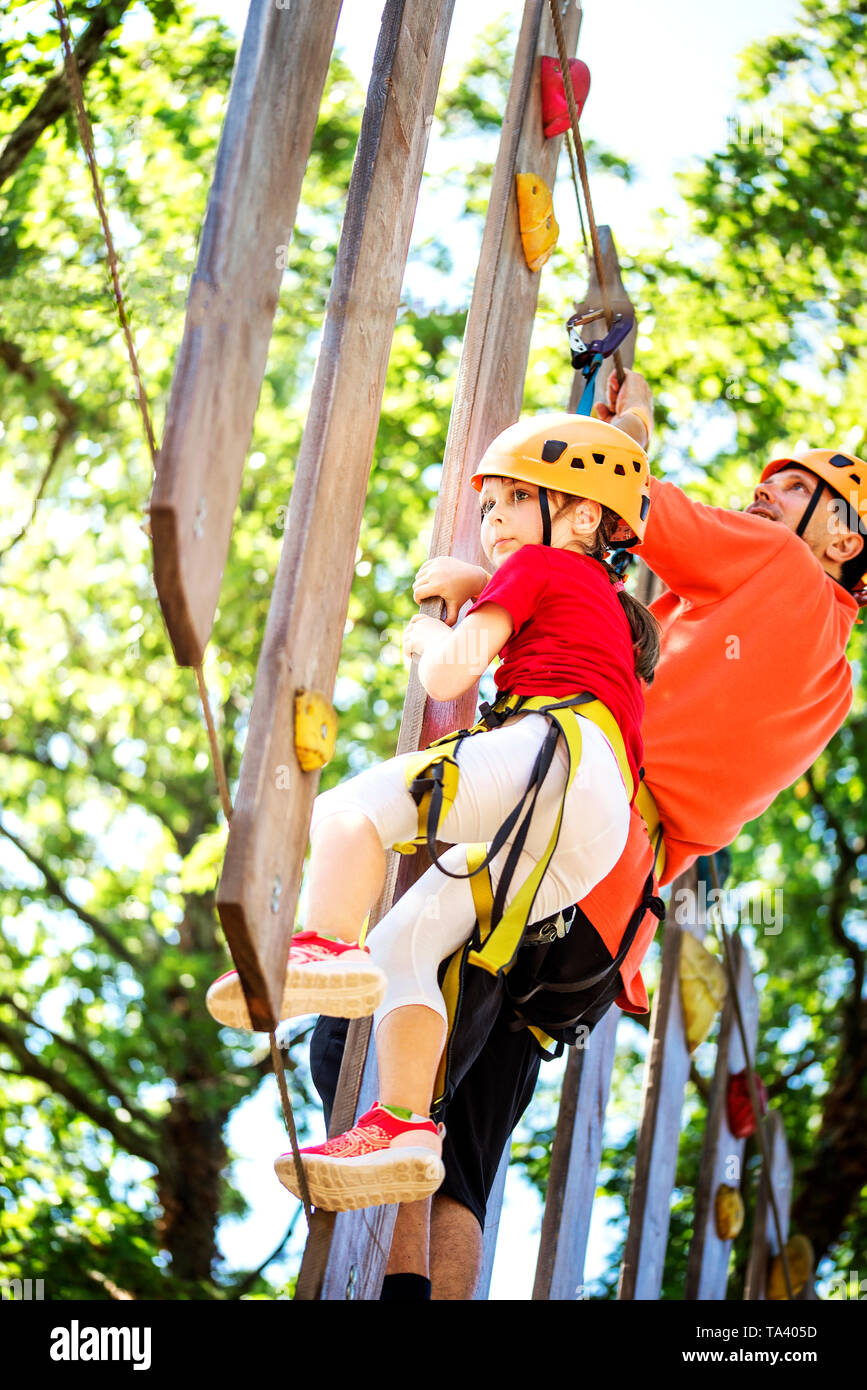 Little brave caucasian girl at outdoor treetop climbing adventure park. 7 years old girlie overcome obstacles on the rope path in the air Stock Photo