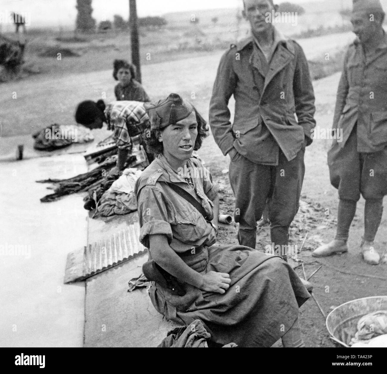 Photo of a Republican female fighter washing clothes during the Battle of Talavera de la Reina behind the front of El Bravo-Orteria. The female soldier is wearing a skirt and a field blouse. In the background, a washboard, soldiers in uniform and women at the washing trough. Stock Photo