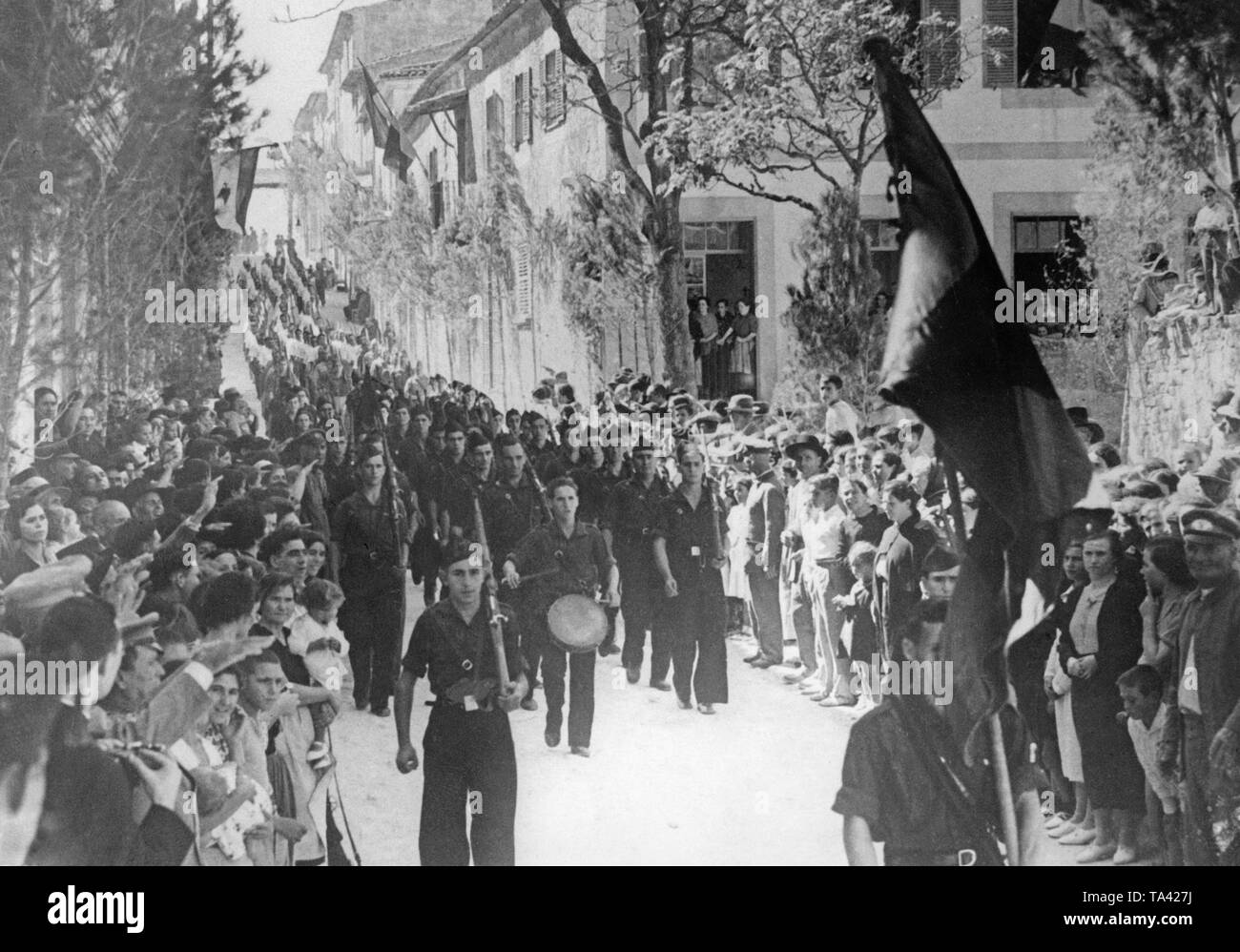 A group of members of the Falange Espanola (Fascist Party of Spain) during a march through Seville, which was conquered by Spanish national forces. The parading soldiers wear the blue shirts of the Falange, and they are armed with carabiners. At the front a drummer marches. In the front, a flag carrier with the red-black-red flag (with yoke and arrows) of the Falange. The inhabitants give the Fascist salute from the pavement. In the background, the Bandera (the flag of Spain) is blowing. Stock Photo