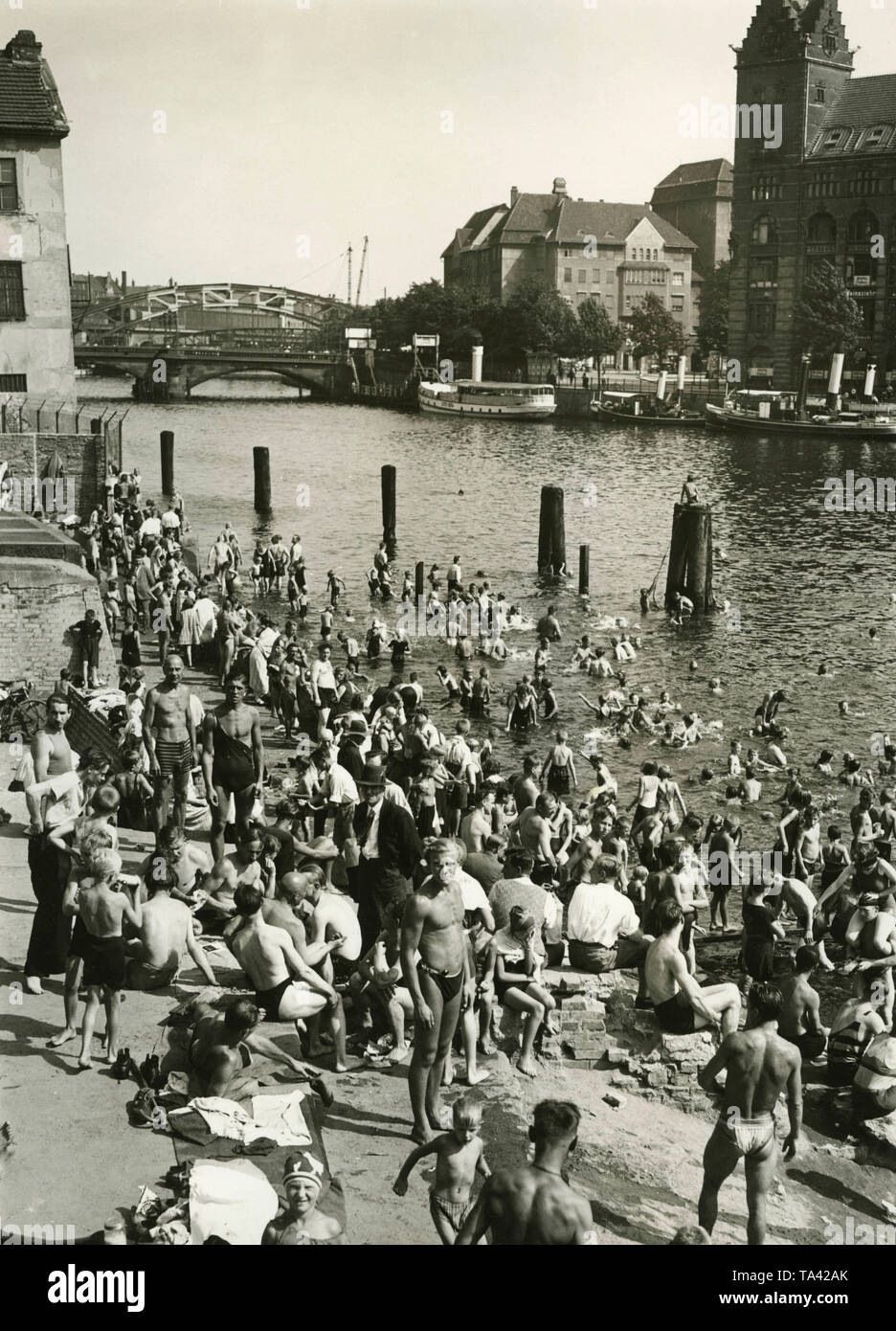 Men and women bathing and sunbathing on the Spree on the side of Stralauerallee (formerly Klein Stralauer Strasse). In the background is the Elsen bridge and a railway bridge. Since the citizens of Berlin can no longer afford to go to the open-air swimming pools, they bathe in the Spree. Stock Photo