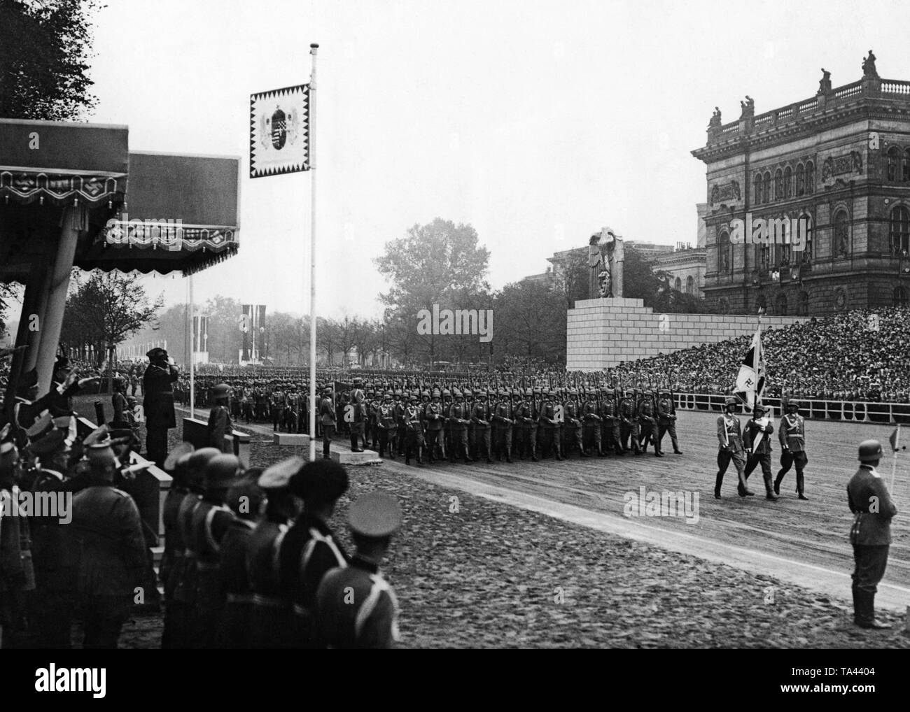 Infantry of the Wehrmacht during a parade along the East-West axis marching past the tribune on which Hitler and the Hungarian head of state Admiral Miklos Horthy (r. saluting) have taken place. The flag is the troop flag of the infantry of the Wehrmacht. The standard next to the grandstand shows the coat of arms of the 'Lands of the Crown of Saint Stephen', including areas lost after 1918. In the background, the Technical University of Berlin. Stock Photo