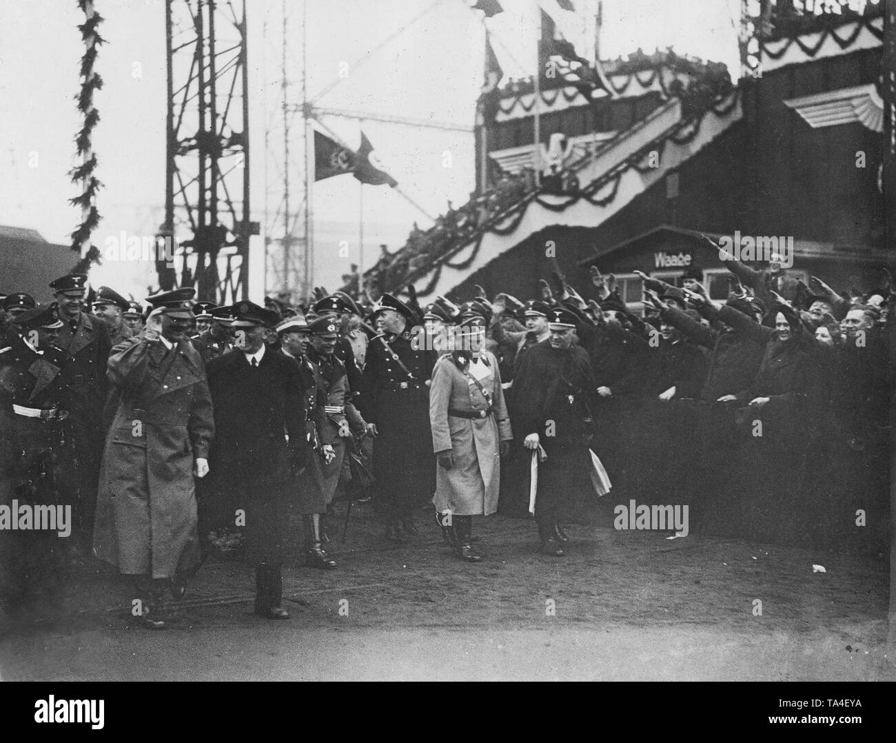 Adolf Hitler (left with his hand raised) arrives at the site of Howaldtswerke Hamburg for the launch of the ship. He is accompanied by the Reichsorganisationsleiter and the name giver of the ship, Robert Ley, as well as SS-Obergruppenfuehrer Josef 'Sepp' Dietrich (both at right in the picture). In the background, a decorated tribune, in front of it a crowd greets the Fuehrer. Stock Photo
