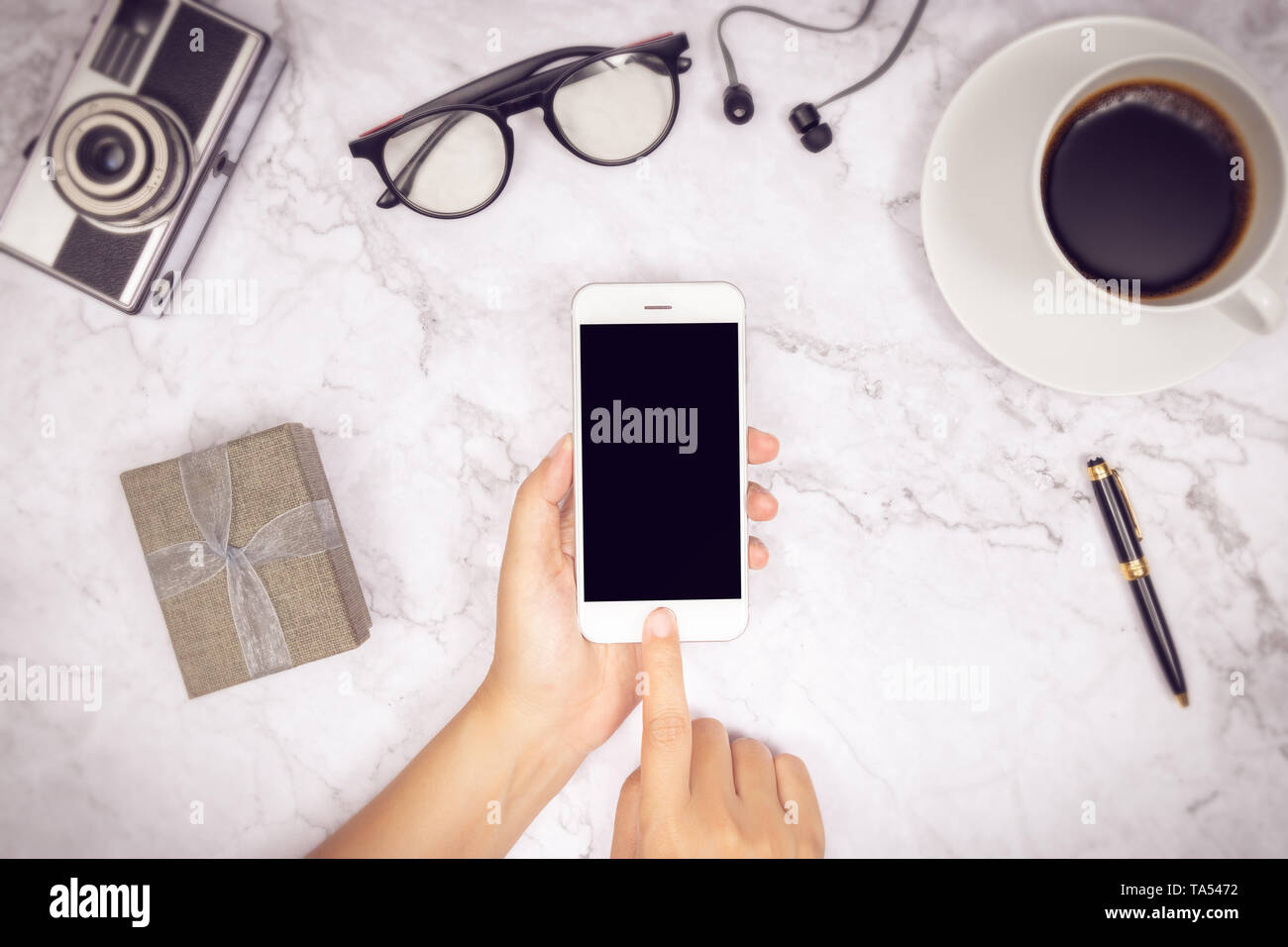 woman hand use mock up of mobile phone blank black screen with finger on touch screen with earphone,  pen, camera, glasses , cup of coffee and gift bo Stock Photo