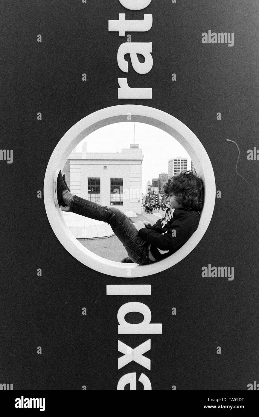 A boy with curly hair and jeans sitting inside the O opening of the Exploratorium sign in San Francisco, California. Stock Photo