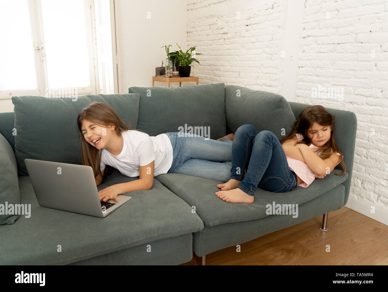 Digital technology addicted young girl playing on the internet using laptop ignoring her sad younger sister. Little girl feeling abandoned and unhappy Stock Photo