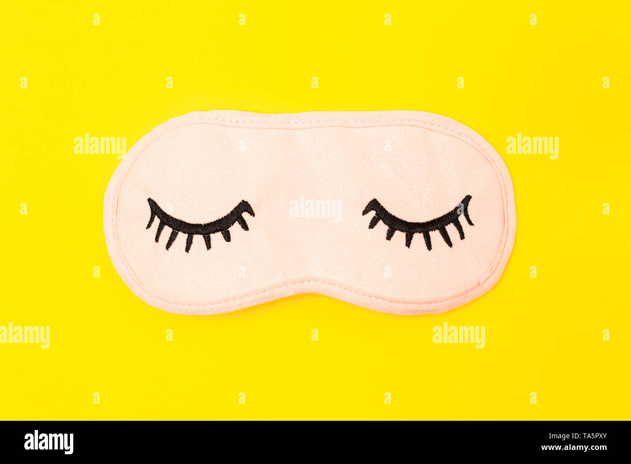 Pastel pink sleep mask with closed eyes embroidered on it with eyelashes on bright yellow neon background. Fashion accessory for sleep. Stock Photo