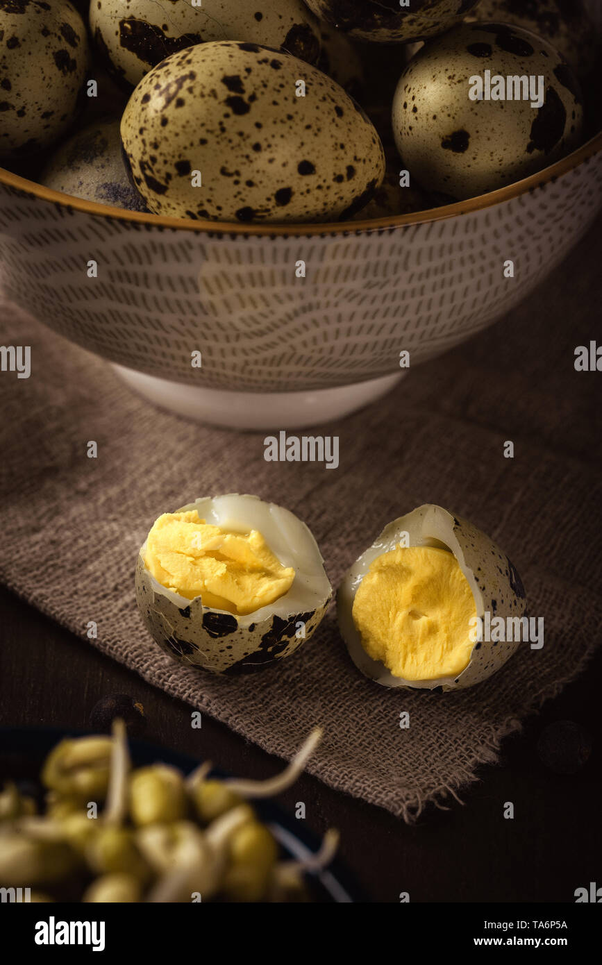 Vertical photo of single boiled quail egg broken in the middle. Other quail eggs are in bowl in background. eggs are placed on wooden board with mung  Stock Photo