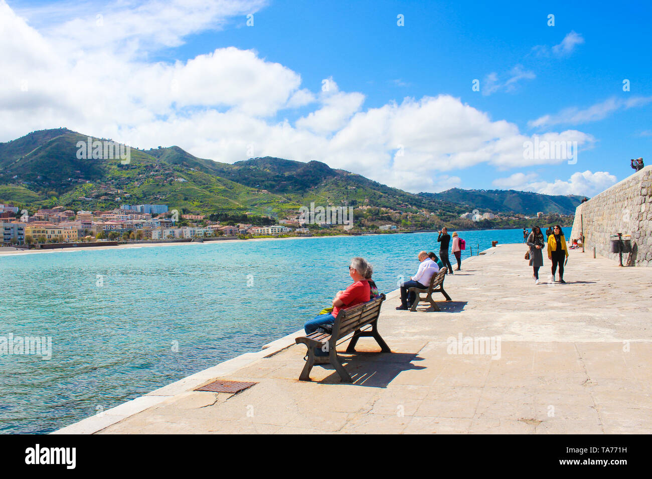 Cefalu, Sicily, Italy - April 7th 2019: People relaxing on the pier on Tyrrhenian coast in Sicilian city. Taken on a sunny day with blue sky. Tourists spending free time. Stock Photo