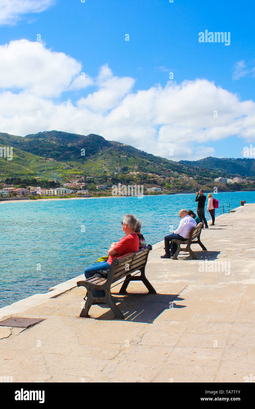 Cefalu, Sicily, Italy - April 7th 2019: People on the pier in beautiful Sicilian city. Tourists spending free time relaxing on the benches. Popular tourist destination. Stock Photo