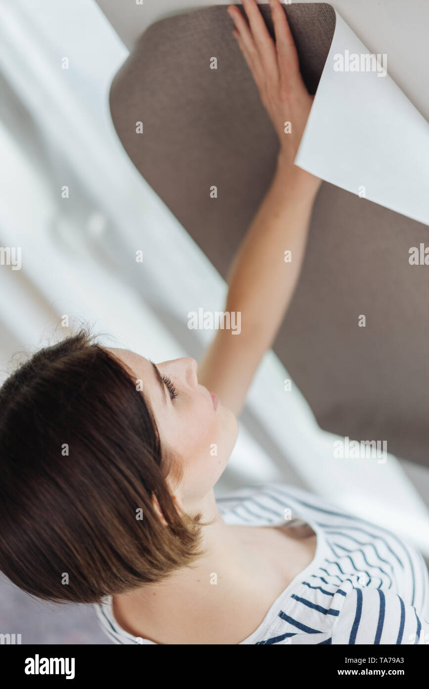 Young woman looking at a wallpaper sample holding it up on a wall in a close up cropped view as she redecorates her home Stock Photo